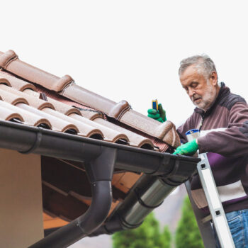 elderly man stands on a metal staircase and repairs a tiled roof against a cloudy sky.