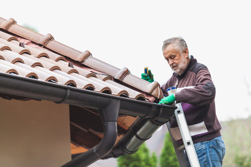 elderly man stands on a metal staircase and repairs a tiled roof against a cloudy sky.