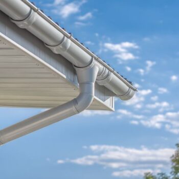 A detailed view showcases a house eave with a white surface and a connected metallic gutter system. The gutter curves downward, contrasted against a vibrant blue sky with soft white clouds. The image is brightly lit, suitable for general home improvement or architectural-related applications.